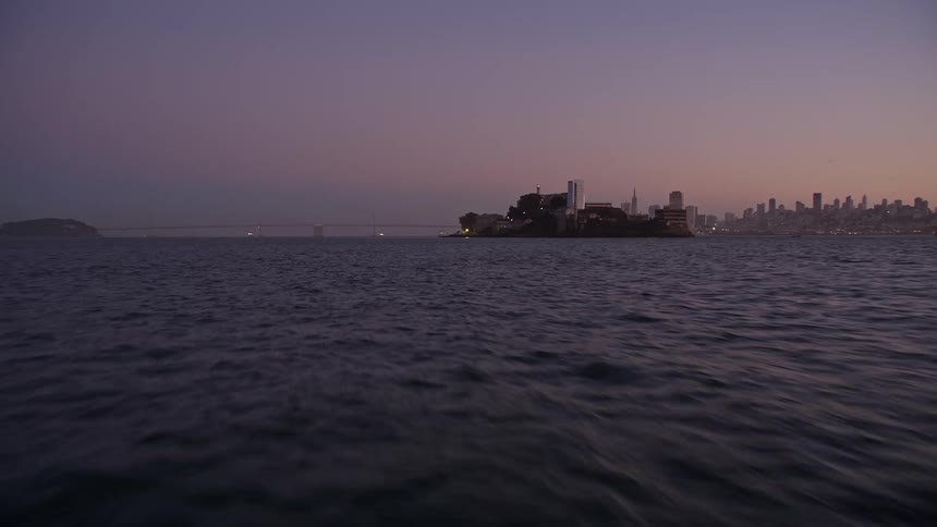 Waves flow past island before passing under bridge leading toward city standing against a pink horizon