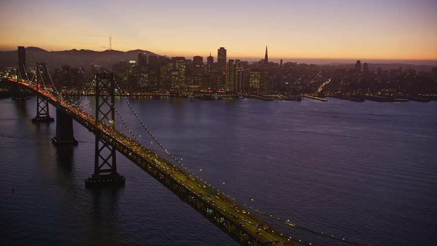 An aerial view of a huge illuminated bridge with several cars passing suspended to the sea and delaying a large city