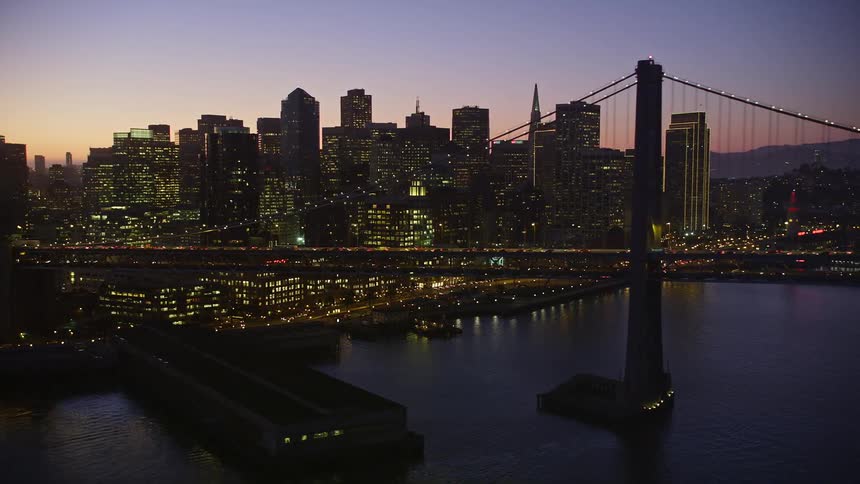 A large suspension bridge in the sea next to a large city that has its lights on at dusk