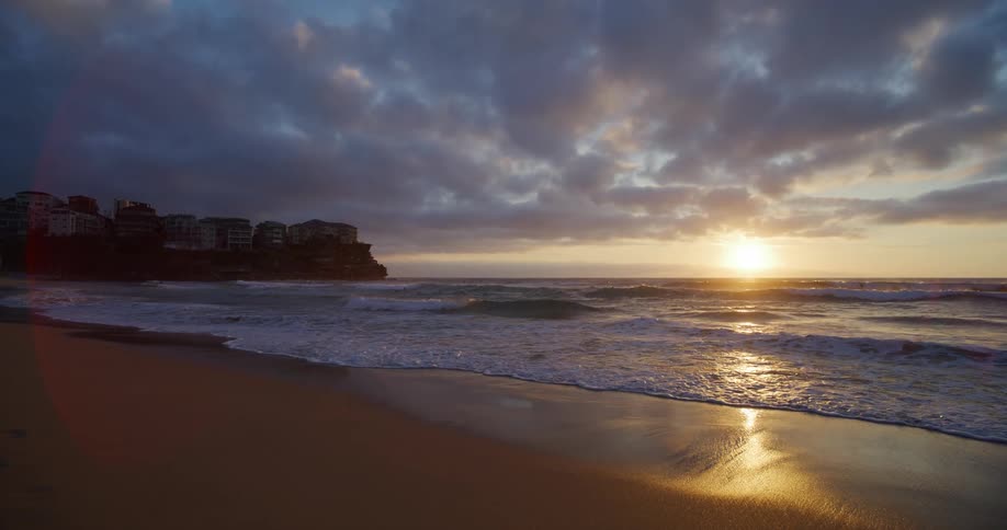 Waves roll over beach sands reflecting the rising sun on an overcast day
