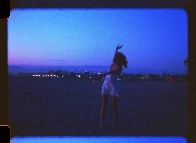 Barefoot woman dances on sandy beach with town in the distance on an evening