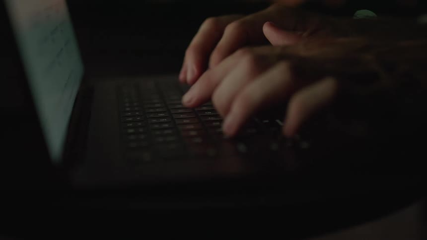 The hands of a person typing quickly on a laptop keyboard with a lit screen
