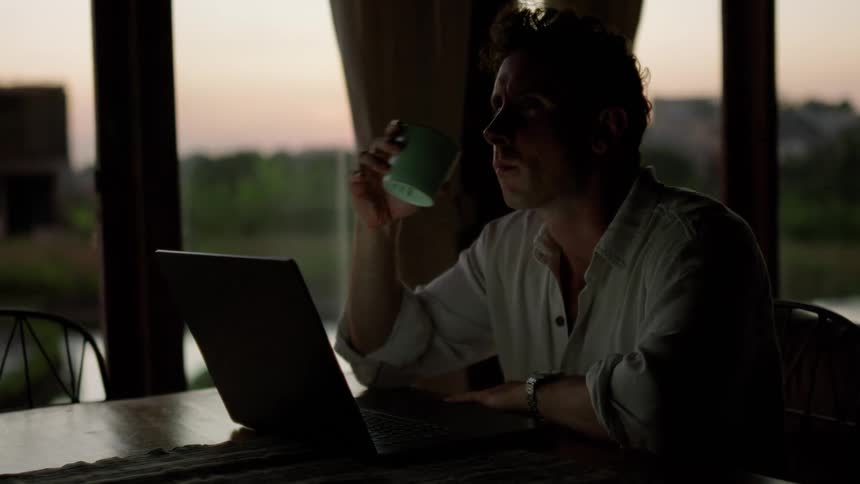 Man sitting at a table with his laptop in front of him and drinking a cup