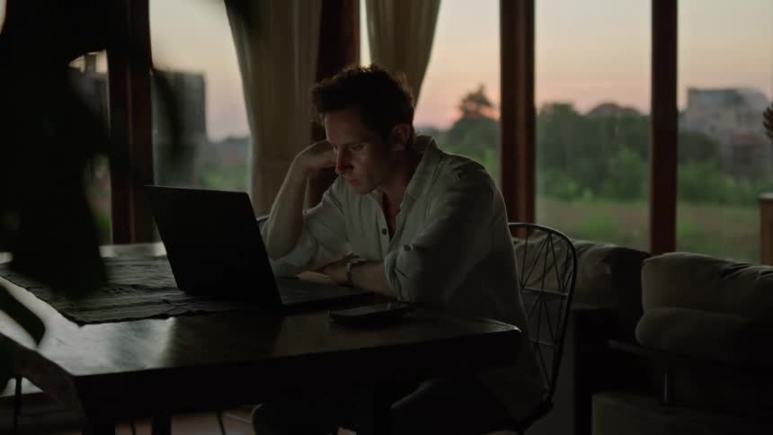 Man wearing a shirt and a watch works on laptop sitting at table in the living room
