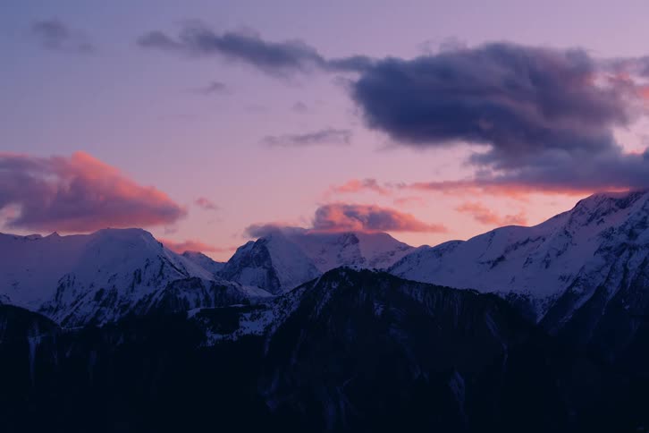 Clouds tinted by the setting sun float over snow covered mountains