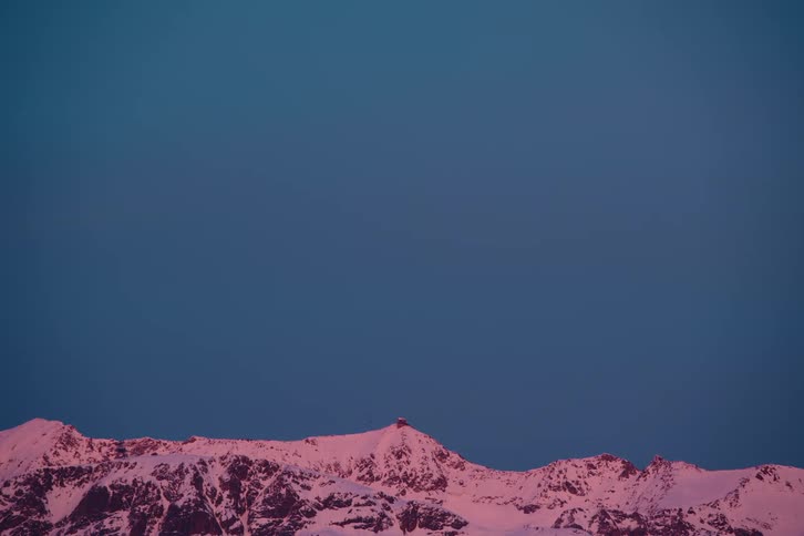 The peak of a snowy mountain in contrast with the clear blue sky and a small black bird flying