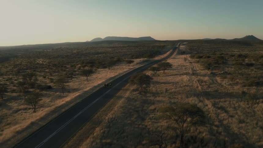 Aerial landscape of a paved highway with two cyclists pedaling and surrounded by desert vegetation with mountains on the horizon on a morning