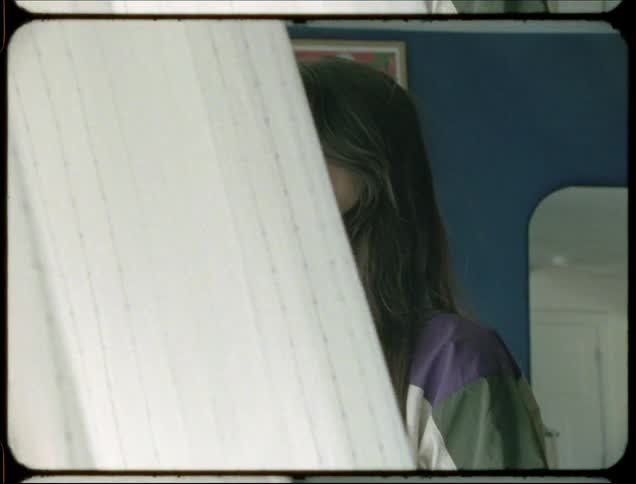 Portrait of a young woman giving a slight smile while standing next to a window with curtains flying beside her