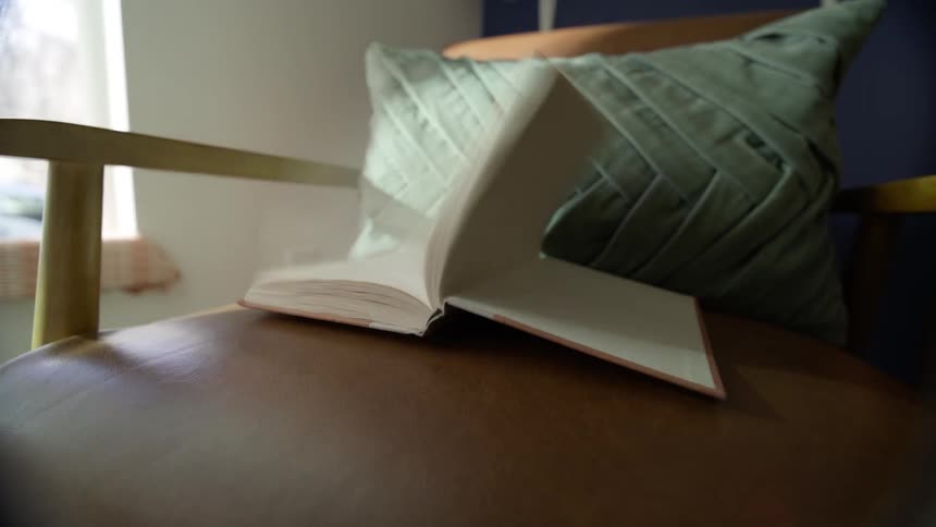 Open book on top of a leather chair that is closed by the force of the wind coming from the window