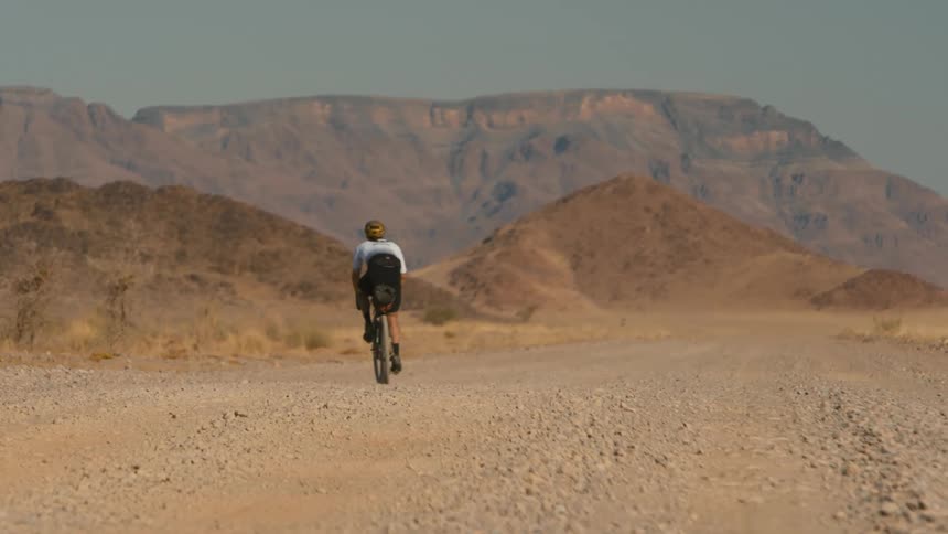 A cyclist is riding on a gravel road in a desert next to the mountains