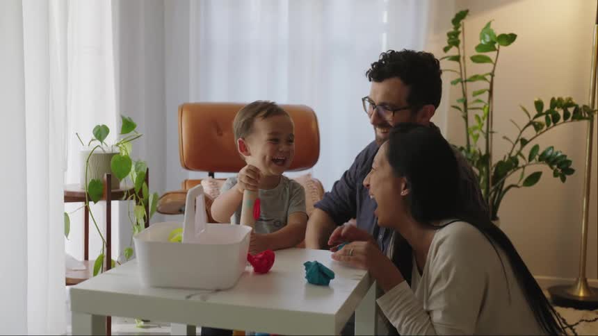 Father and mother smiling a lot next to their little son while they are around a table playing with modeling clay