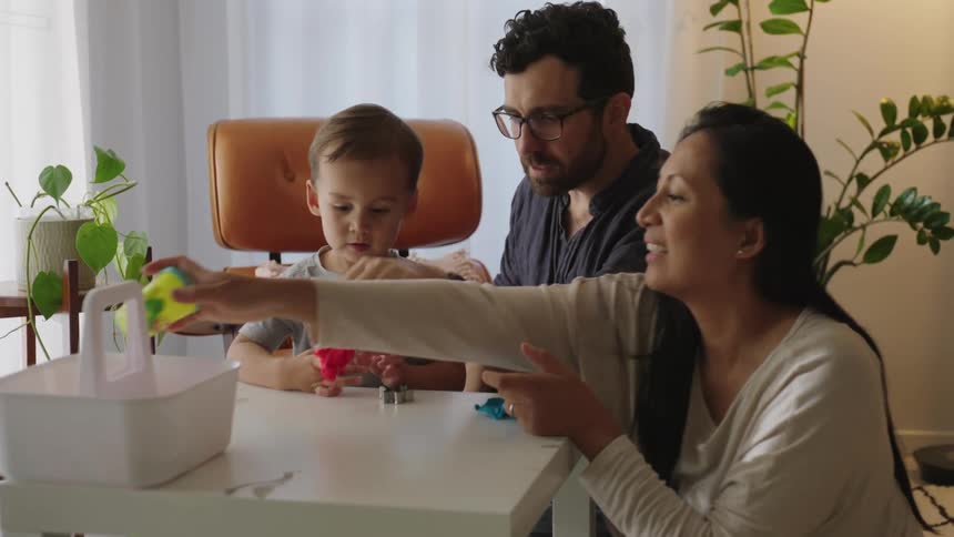 A family of parents and a boy who are around a small white table playing with the child who has toys in his hand