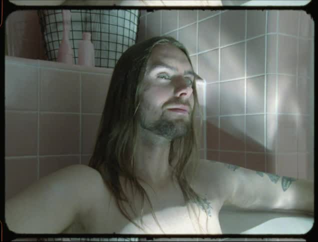 A tattooed man sits back in a water filled bathtub grinning and taking in his surroundings