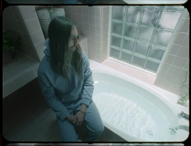 A person wearing a blue hoodie and pants is seated on the edge of a white bathtub