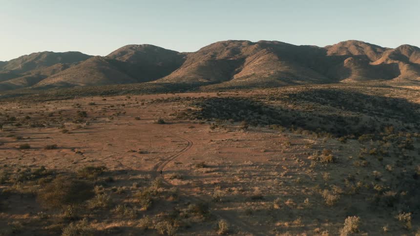 Aerial scene of a person riding a bicycle in a natural trail environment with mountains on the horizon on a blue sky morning