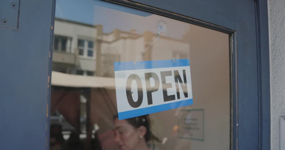 A young woman changes the side of a sign that says closed to open and the sign is hanging on a glass door
