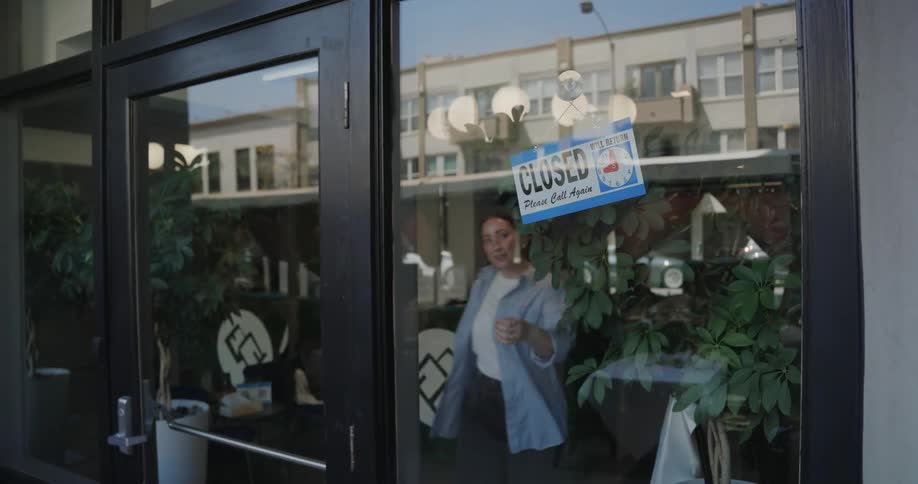 A female small business owner flips the sign on her shop door from "Closed" to "Open"