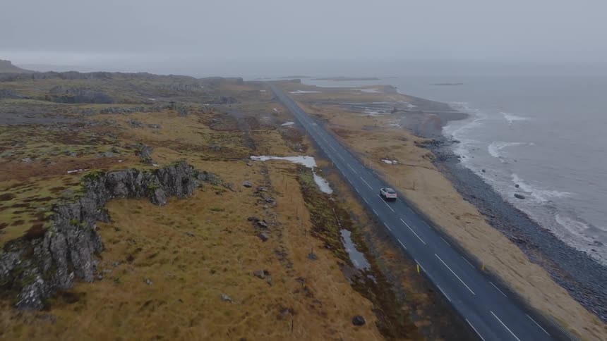 An aerial view of a car driving on an asphalt road next to the sea coast