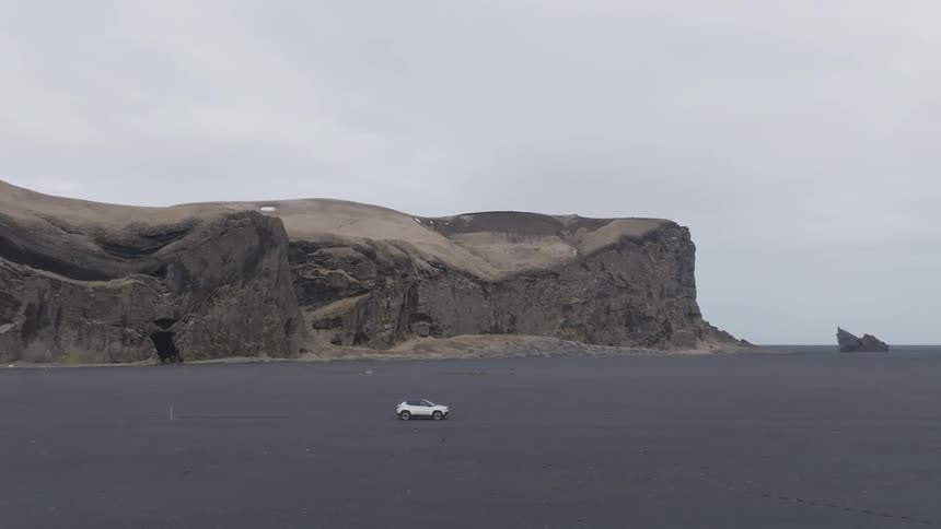 A white car driving in a region with black earth and a stone mountain in the background