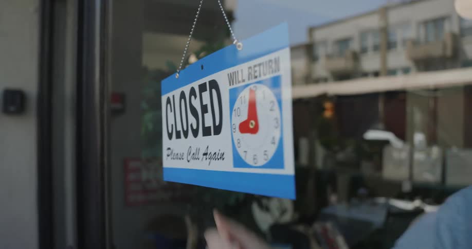 A human hand changing a sign that says closed to open and is hanging on a glass door