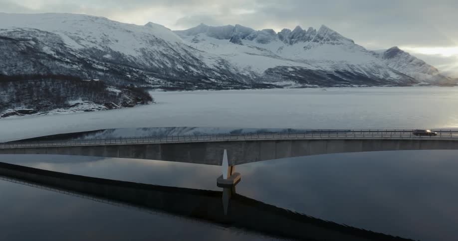 Aerial panorama of a bridge over a lake with vehicle traffic and surrounding sunset on the horizon and mountains covered in snow