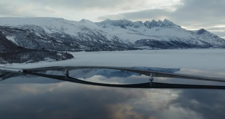 Aerial panorama of a bridge over a lake surrounded by snowy mountains on a cloudy morning