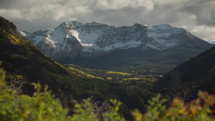 Deciduous and conifer trees fill valley and mountains bordering snowy mountain on a cloudy day