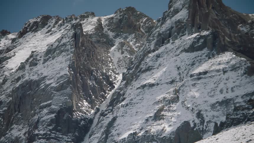 A rugged mountain slope and peak with patches of snow under a blue sky