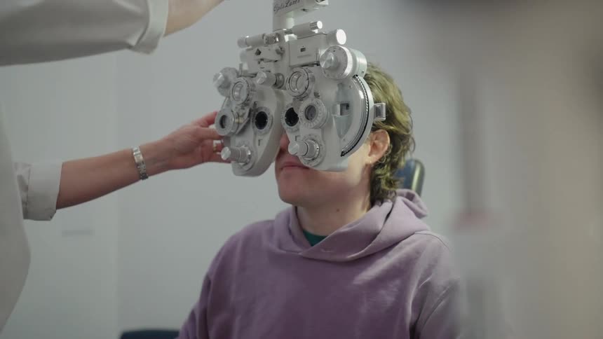 A seated woman is undergoing an eye exam with a phoropter while a healthcare professional adjusts the equipment