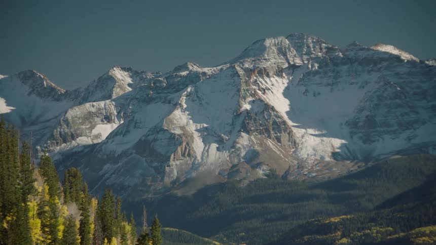Landscape of a hill with conifers and surrounding mountains with cliffs covered in snow