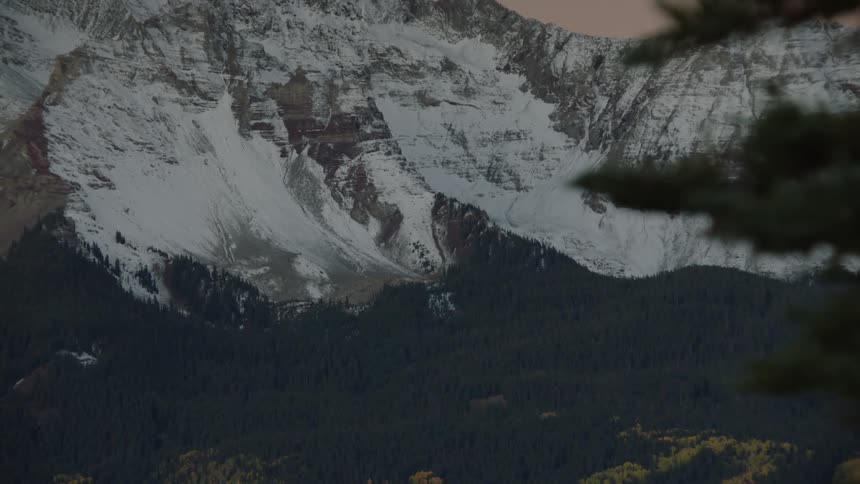 Deciduous forest borders conifer forest at the base of snowy mountains under a red tinted sky