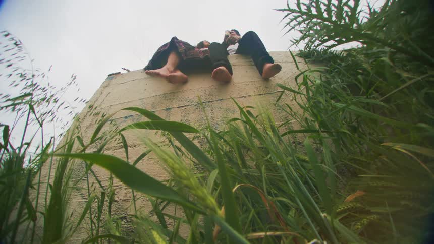 A barefoot couple sitting atop a large concrete wall surrounded by tall grass