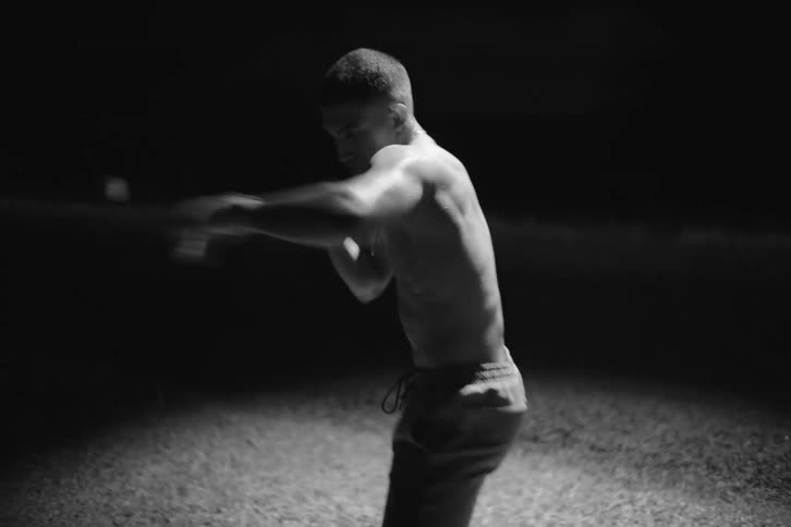 A young shirtless boxer practices under a spotlight near a street at night