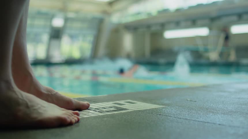 Feet of a person next to a swimming pool with sports tracks and people swimming