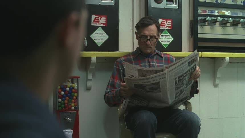 Side of a mans face in the foreground and in the background another man sitting reading a newspaper