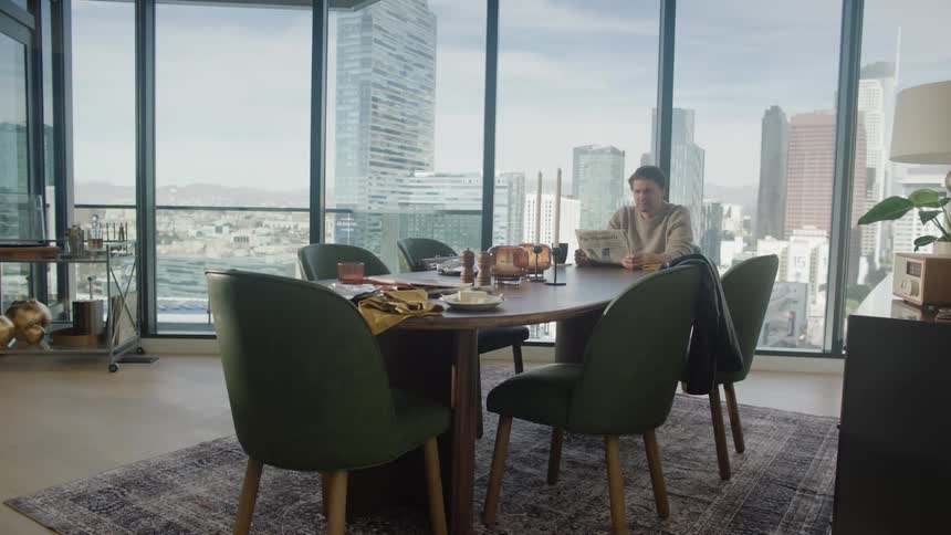 A man sitting alone at a meal table in a tall building reads a newspaper in front of a large window with a view of a block of skyscrapers