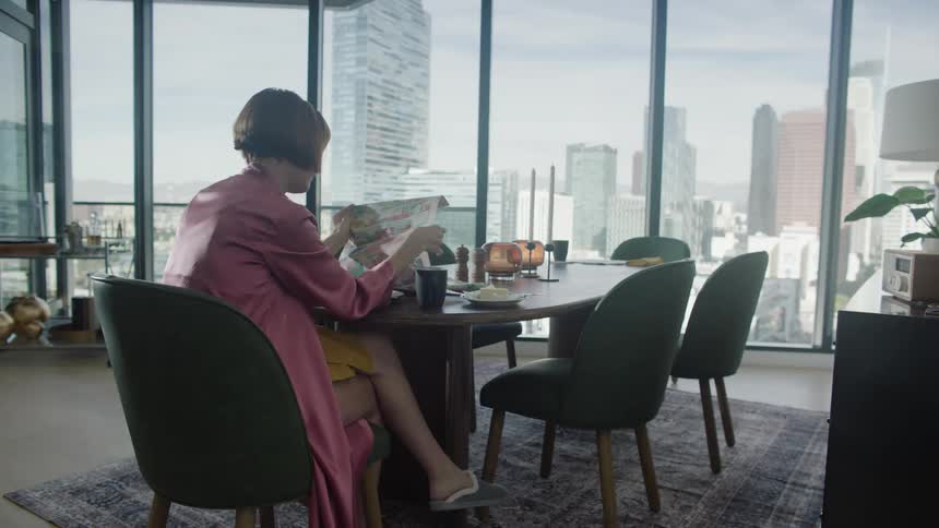 A woman seated at a dining table reading a newspaper with a cityscape visible through large windows