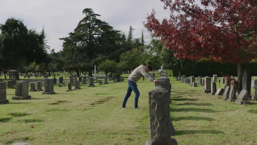 A man visits a grave and walks up to the headstone he leans on it