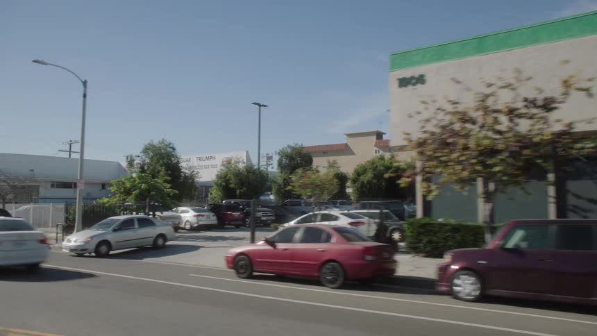 A vehicle is driving down a highway near buildings passing by cars parked near the road