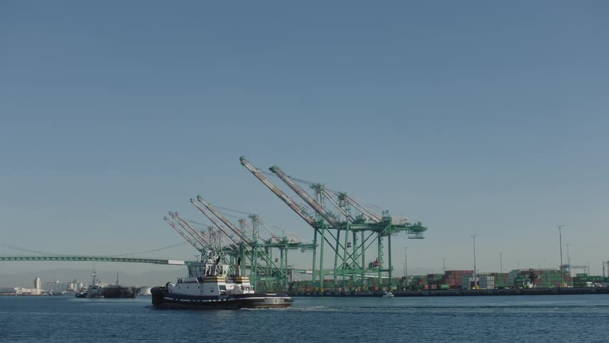 A boat on a river passes in front of cargo cranes from a dock