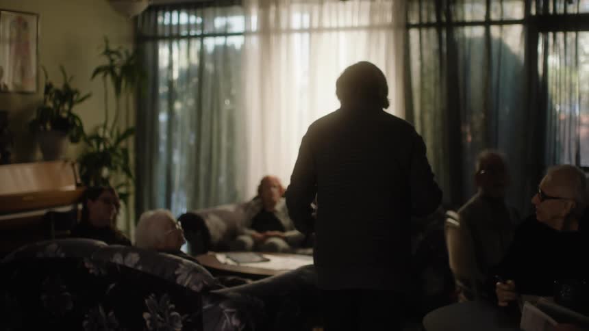 Man standing talking to elderly people who are sitting at tables and armchairs around a room