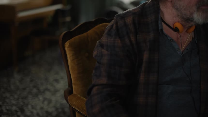 A man with a gray beard and a headset around his neck is sitting in a chair