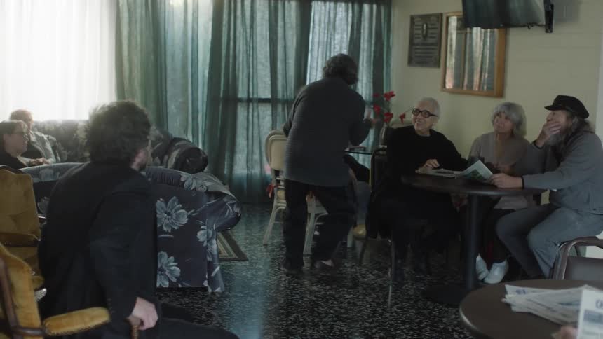 Group of people sitting at a table with a man standing passing by the tables and gesturing his arms talking