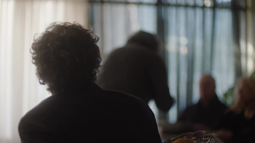 Man sitting at a table watching another person in the background talking to elderly people at another table