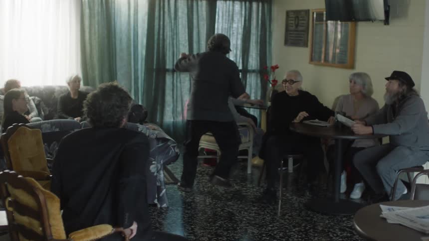 Elderly people sitting at a table and in armchairs with a man playing and talking to them