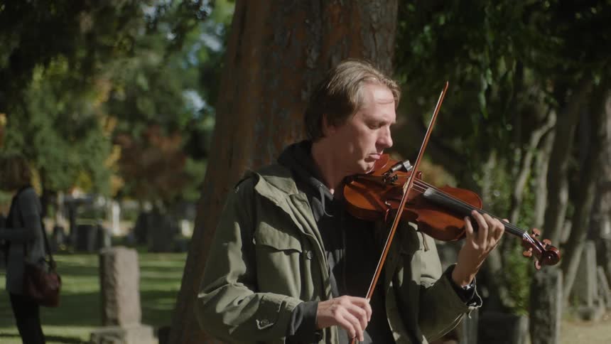 A man is playing the violin in a cemetery and behind him there is a tree