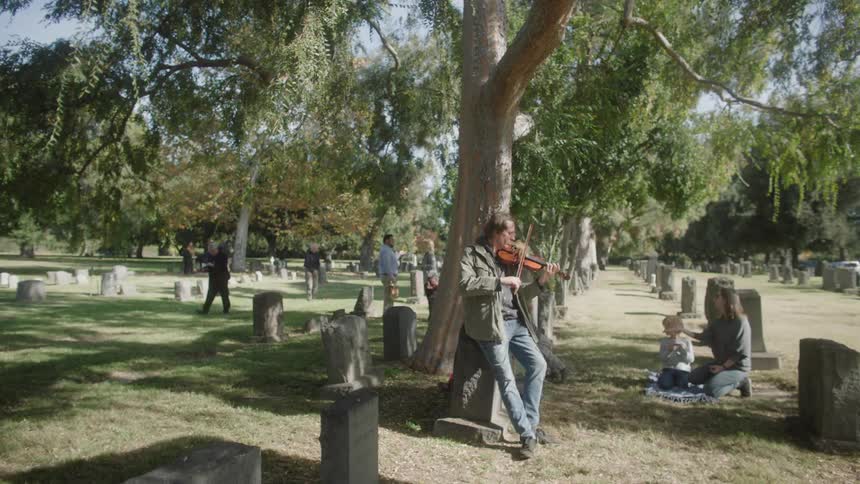 A man plays the violin while leaning on a grave