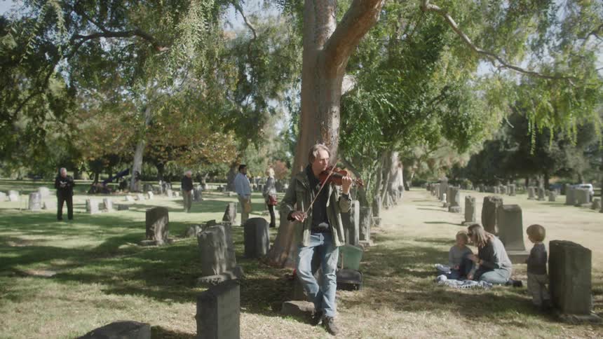 A man plays violin in a graveyard and a boy nearby approaches him and says hello