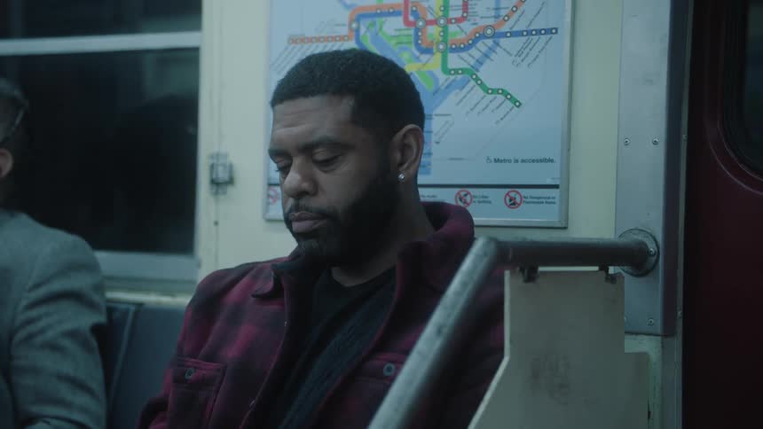 A man in a red jacket is sitting on a subway seat and is looking down