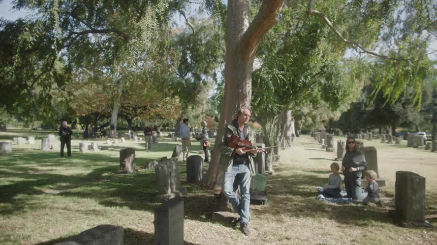 Man leaning against a tree playing a violin in the middle of a cemetery with people around